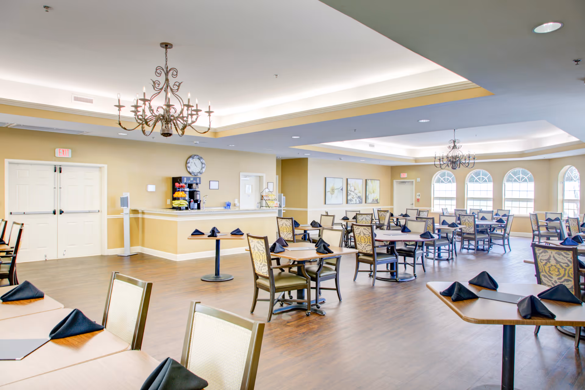 A spacious dining room with multiple tables and chairs arranged neatly. Each table has black folded napkins placed on it. The room features large arched windows allowing natural light to fill the space, two decorative chandeliers hanging from the ceiling, and a counter area with a coffee machine and fruit display against a beige wall.