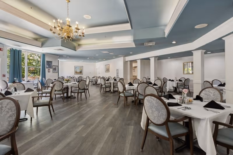 A spacious dining room with multiple round tables covered with white tablecloths, each set with black napkins, silverware, and small flower arrangements. The room has large windows with blue curtains, a chandelier hanging from the ceiling, and light-colored walls with framed artwork.