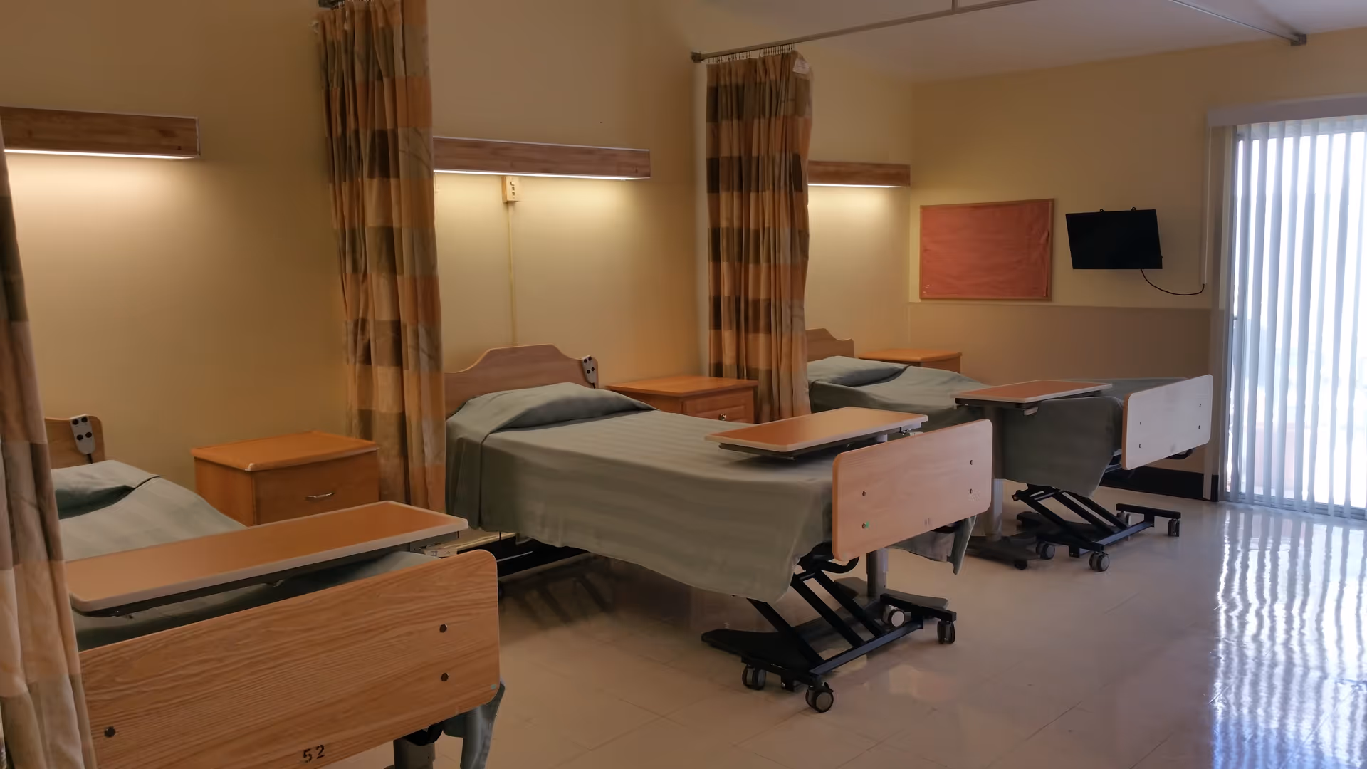 A room with three hospital-style beds arranged in a row, each with a greenish bedspread and an overbed table. There are privacy curtains between the beds, wooden nightstands next to each bed, and a small wall-mounted TV on the far wall. The room has a large window with vertical blinds allowing natural light to enter.