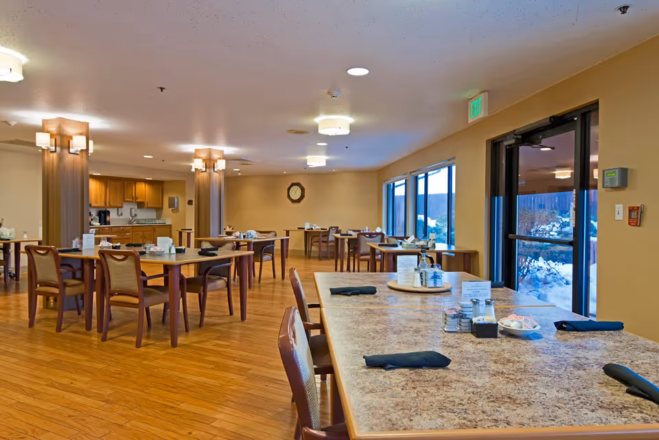 A spacious dining room in Highland Care Center with multiple wooden tables and chairs arranged neatly. The tables are set with black napkins, condiments, and menus. Large windows on one side provide natural light and a view of a snowy outdoor area. The room has warm lighting and wooden flooring, with a clock on the far wall and an exit door near the windows.