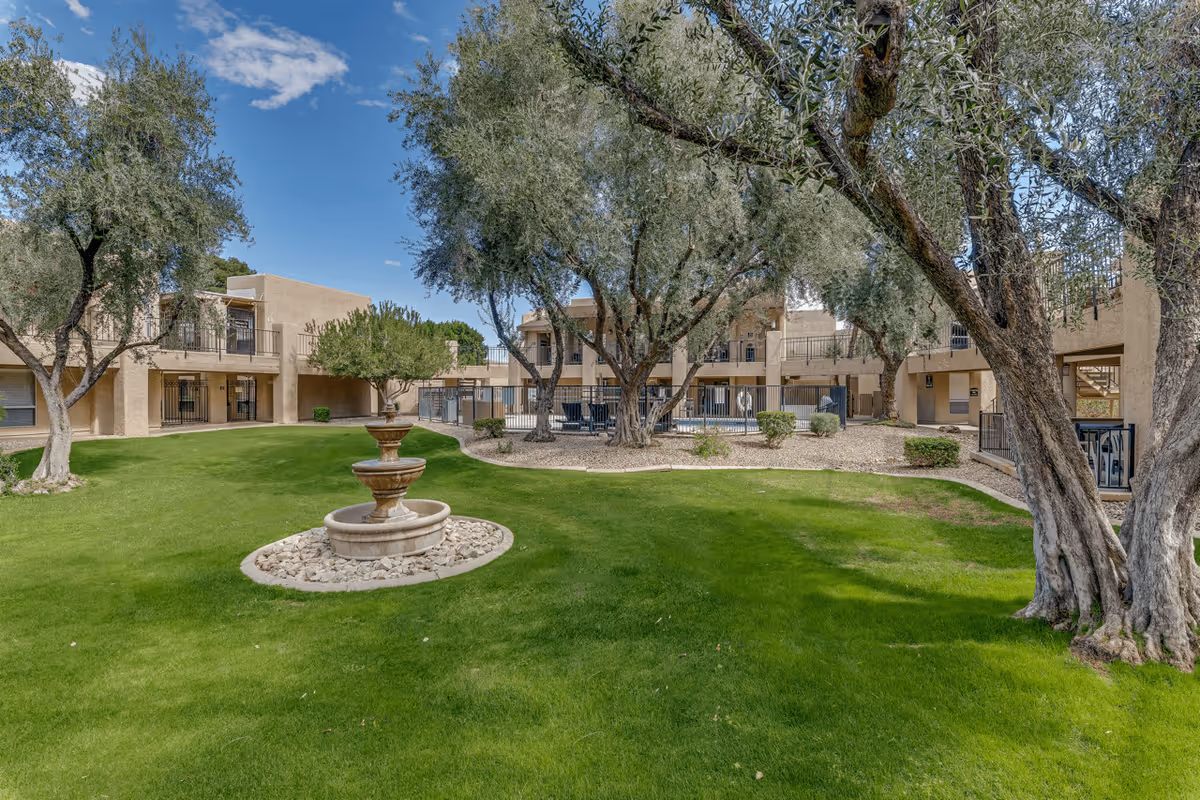 Outdoor courtyard area at Scottsdale Village Square featuring green grass, several mature trees, a stone fountain in the center, and beige two-story buildings surrounding the space under a blue sky with some clouds.