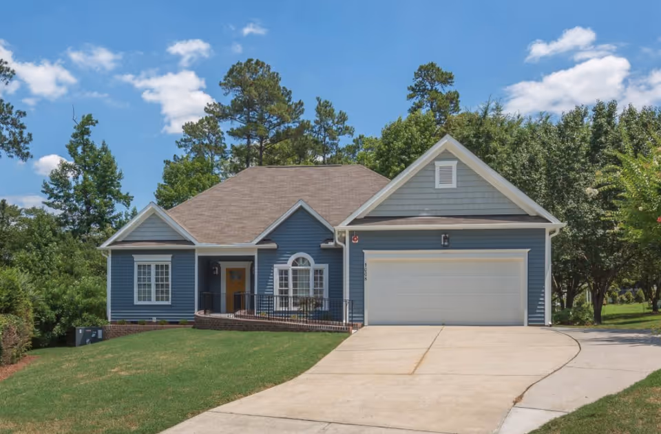Front exterior view of a single-story blue house with a two-car garage, a sloped driveway, and a small front porch with a railing. The house is surrounded by green grass, trees, and bushes under a partly cloudy blue sky.