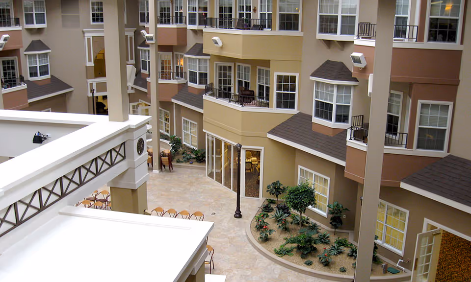 Indoor courtyard area of a senior living facility with multiple floors of apartments featuring balconies and large windows. The courtyard has tiled flooring, a small garden with plants, and several tables and chairs arranged for seating.