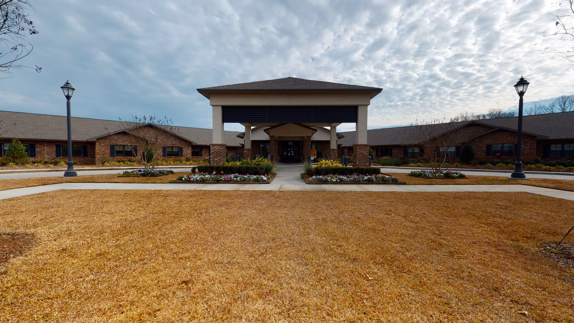 Front exterior view of Landmark Nursing Center Hammond showing a single-story brick building with a covered entrance, surrounded by landscaped flower beds and a large lawn area under a cloudy sky.