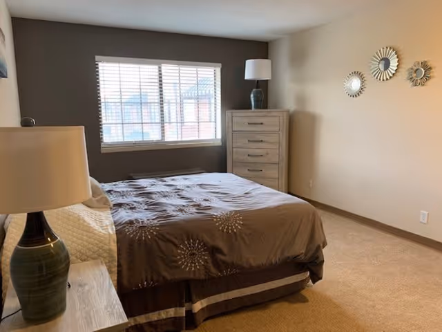 Well-lit bedroom with a bed, two lamps, a tall dresser, and a window with blinds.