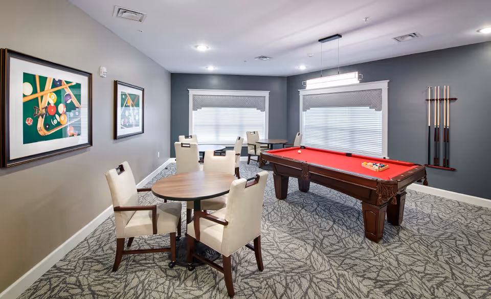A recreational room with a red felt pool table, pool cues mounted on the wall, and several round tables with beige upholstered chairs. The room has gray walls, two large windows with blinds, and framed pictures of billiard balls and cues on one wall.
