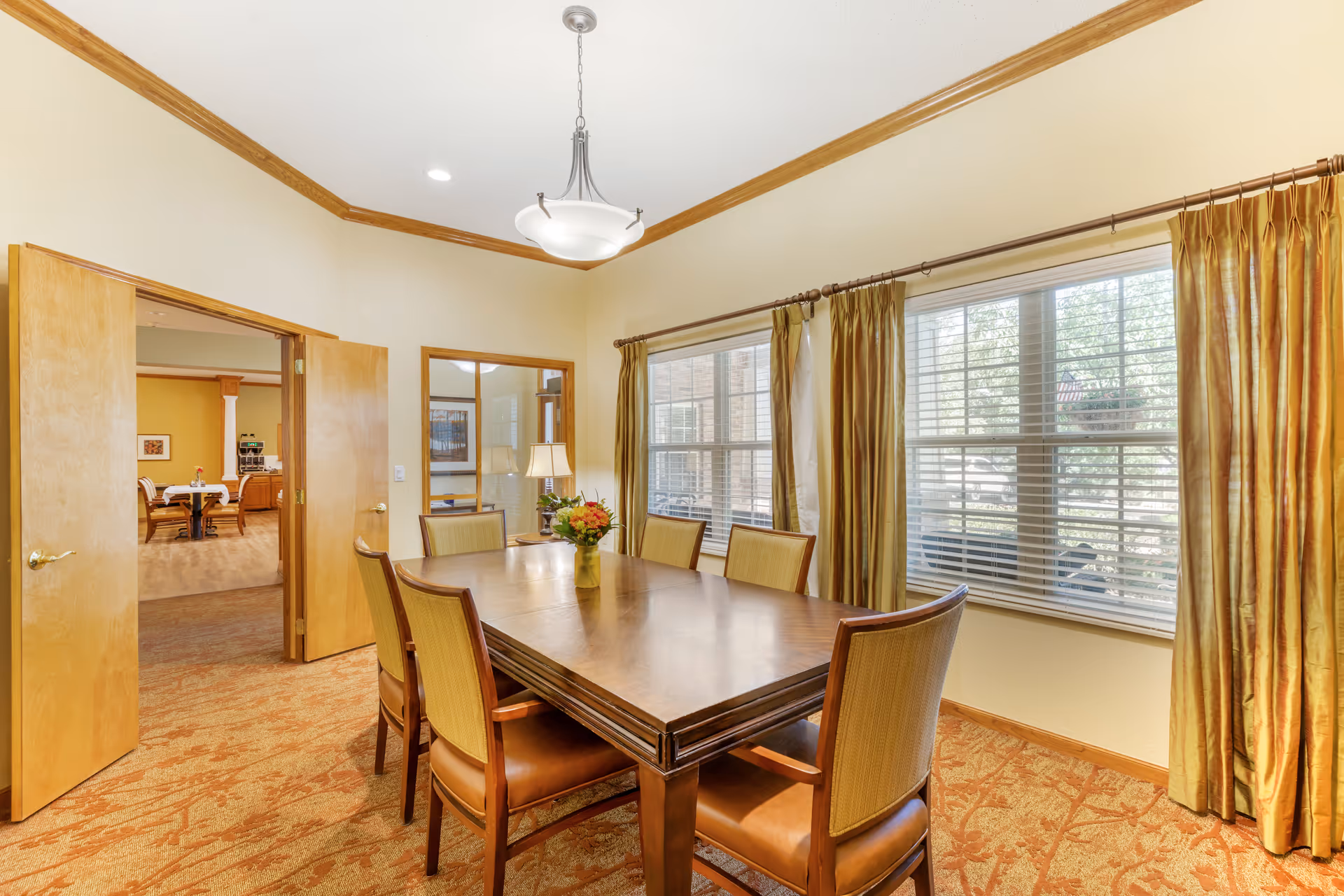 A well-lit dining room with a rectangular wooden table surrounded by six cushioned chairs. The room features large windows with gold curtains, beige walls with wooden trim, and a hanging light fixture above the table. An open doorway leads to another room with additional tables and chairs.