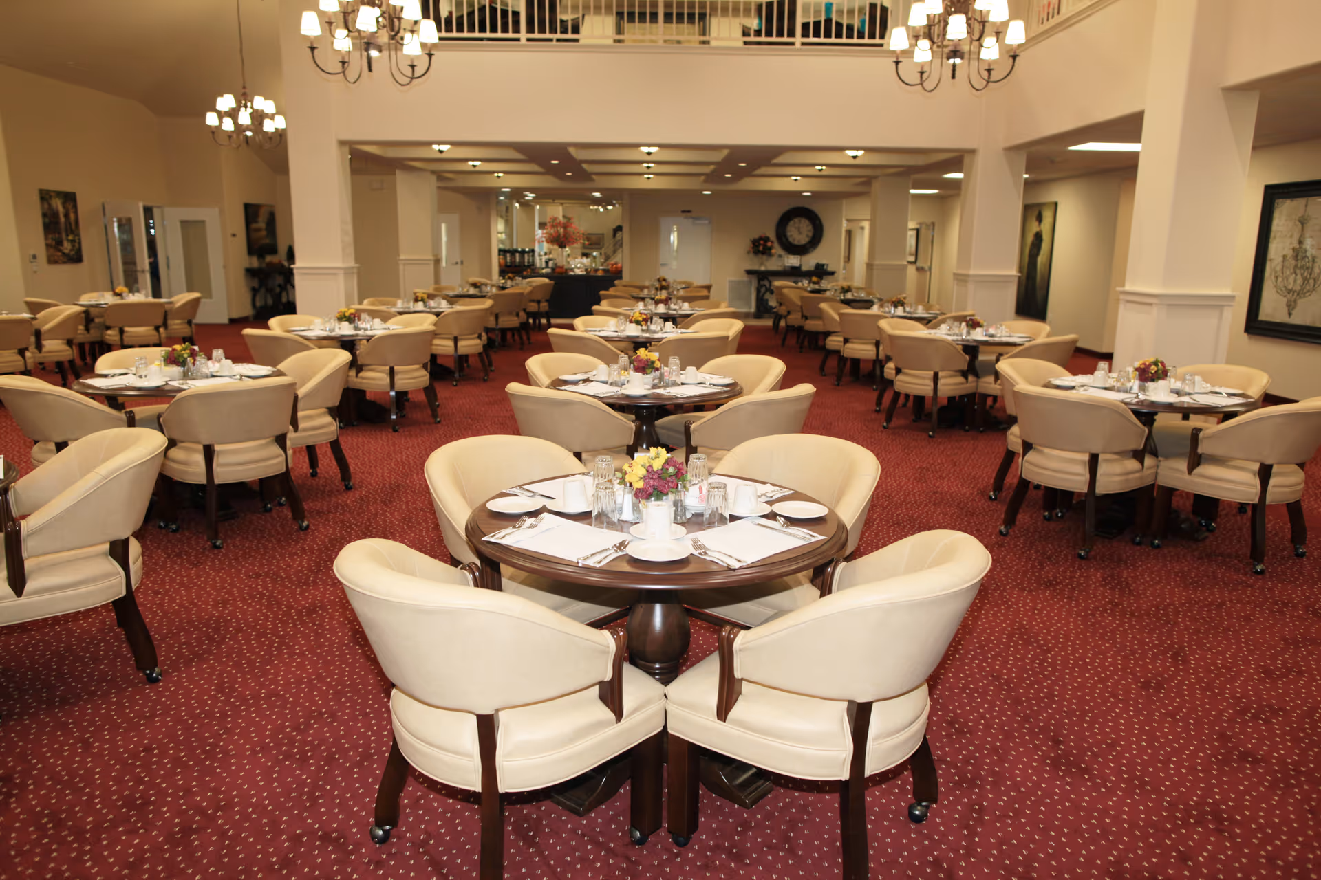 A spacious dining room with multiple round tables set for meals, each surrounded by beige upholstered chairs. The room features red carpet flooring, chandeliers hanging from the ceiling, and decorative wall art. The tables are neatly arranged with white tableware, glasses, and small floral centerpieces.