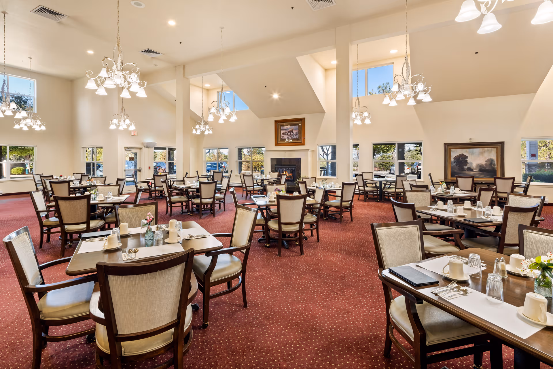 A spacious senior living dining room with multiple tables and chairs arranged neatly. Each table is set with placemats, cups, glasses, and small flower vases. The room has high ceilings with several chandeliers providing warm lighting. Large windows allow natural light to enter, and there are framed paintings on the walls. The carpet is red with a subtle pattern.