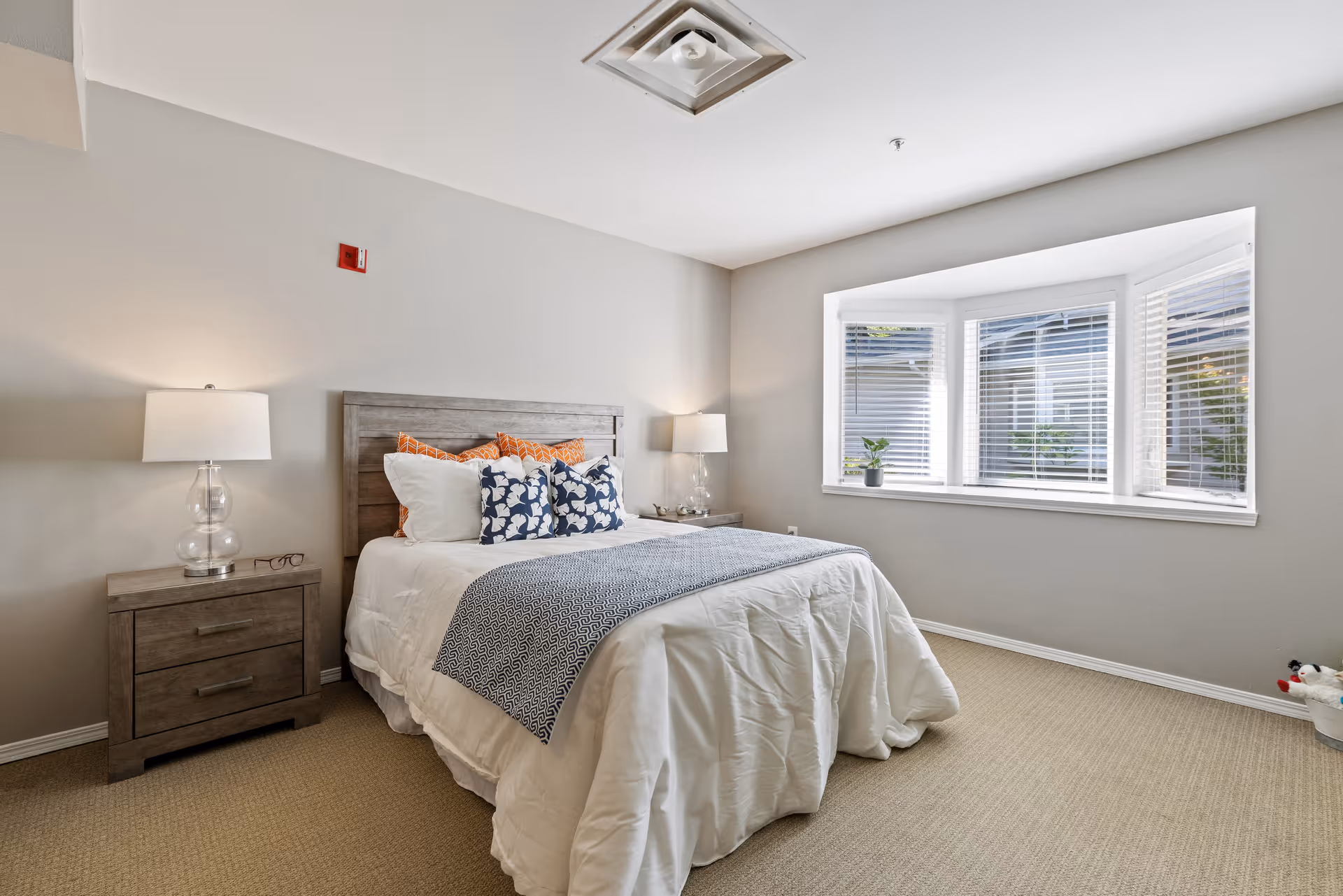 A neatly made bed with white bedding and decorative pillows in a bedroom. The bed has a wooden headboard and is flanked by two wooden nightstands, each with a clear glass lamp. There is a large bay window with white blinds and a small potted plant on the windowsill. The room has beige carpet and light-colored walls.