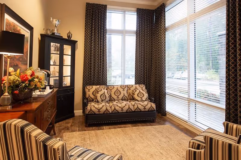 A cozy living room with a patterned loveseat against a window with dark curtains, two striped armchairs, a wooden sideboard with a lamp and flower arrangement, and a black display cabinet with trophies and decorative items.