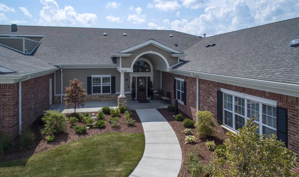 Front exterior view of a single-story senior living facility with a brick facade, a gray shingled roof, a curved concrete walkway leading to the entrance, and landscaped greenery including bushes and small trees under a partly cloudy sky.