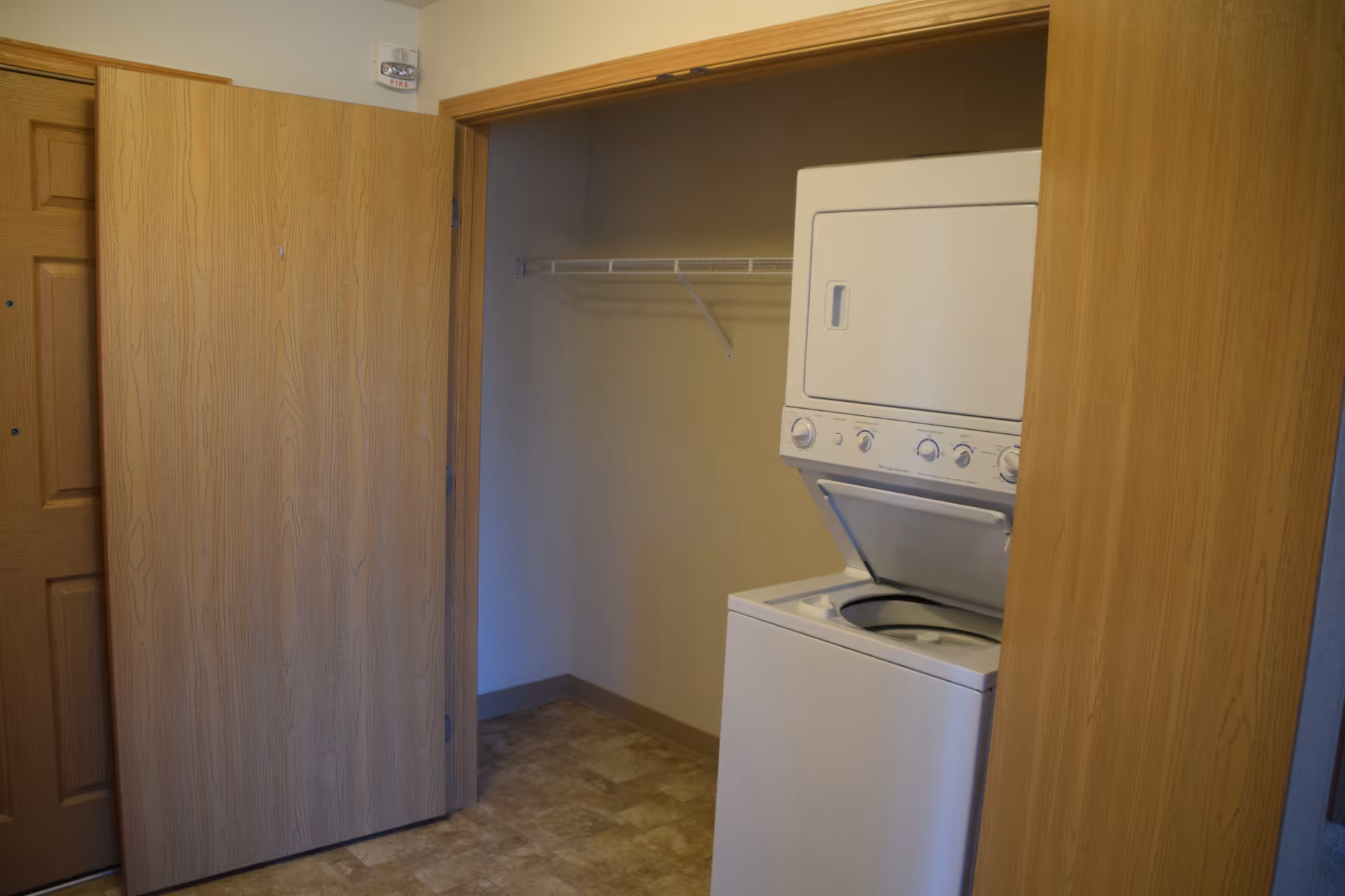 Laundry area with a stacked washer and dryer unit inside a closet with wooden sliding doors partially open. There is a wire shelf above the washer and dryer, and the floor has a beige tile pattern.
