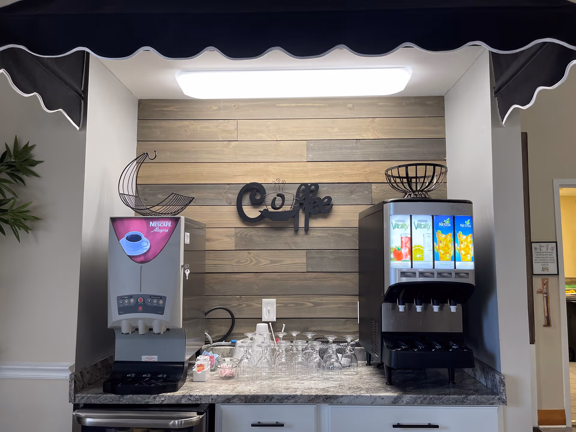 A beverage station with a coffee machine on the left and a juice dispenser on the right, set against a wooden plank wall with a decorative sign that says 'Coffee'. Various glasses and cups are arranged on the countertop below the machines.