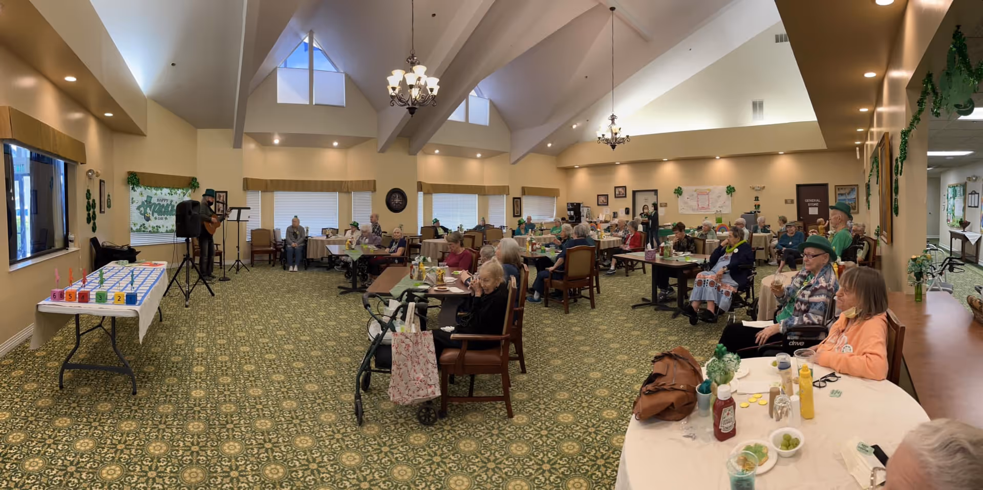 A large room with high vaulted ceilings and chandeliers filled with elderly residents seated at round tables. A man is playing a guitar and singing at the front left side of the room. The room is decorated with green shamrocks and other St. Patrick's Day decorations. There is a table with a game setup on the left side near the musician. The carpet has a green and yellow pattern, and the walls are painted a light beige color.