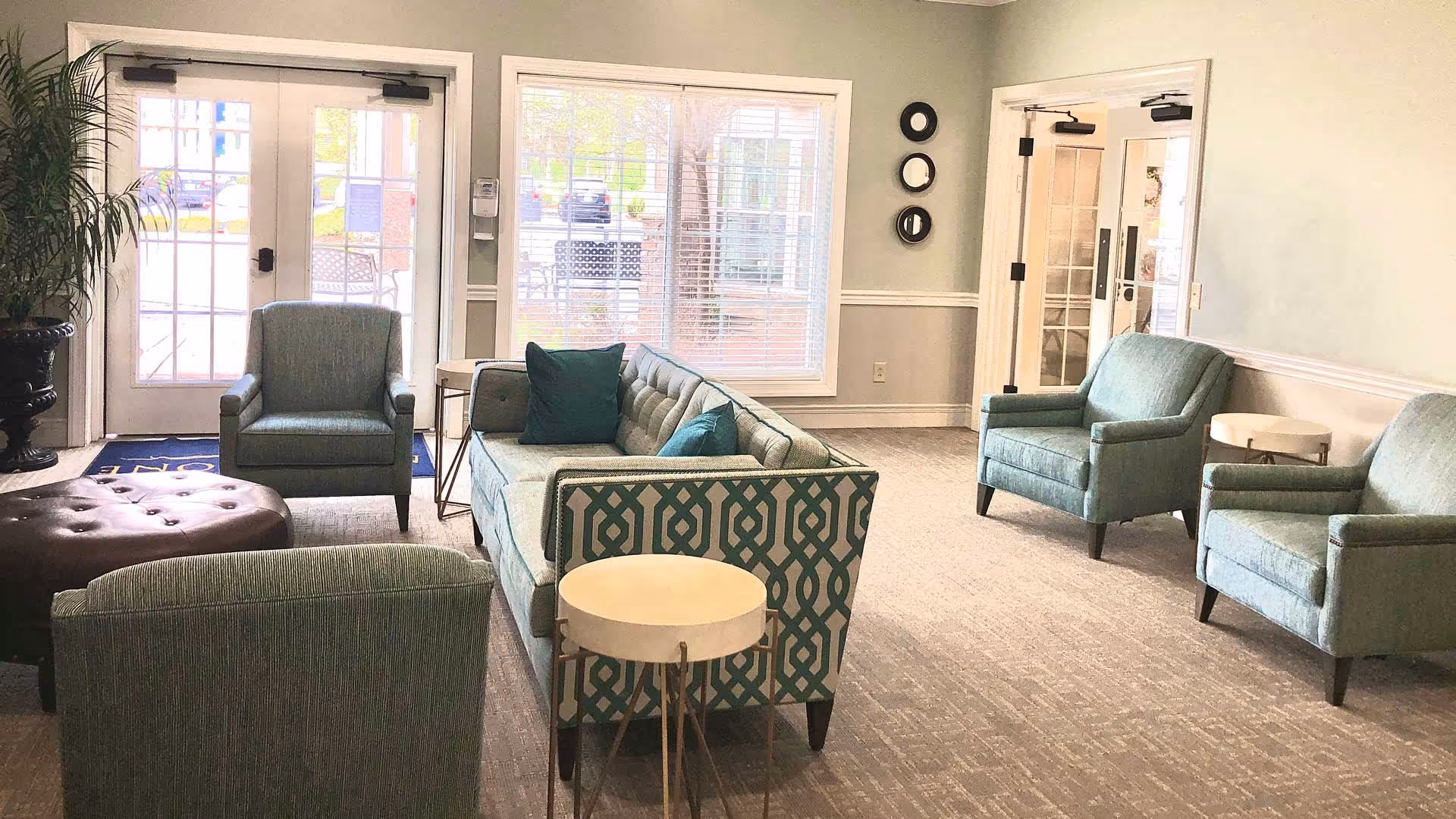 A bright and spacious living room area with light blue upholstered chairs and a patterned sofa arranged around a brown tufted ottoman. There are two small round side tables, large windows letting in natural light, and glass doors leading outside. The walls are painted light green with white trim, and there are three decorative round mirrors on one wall.