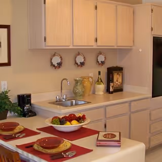 A kitchen area with light wood cabinets, a small sink, and a countertop. On the counter are decorative vases, a bottle of wine, and a coffee maker. In the foreground, a dining counter is set with two place settings including plates, bowls, utensils, and red placemats. A bowl of fruit is also on the dining counter.