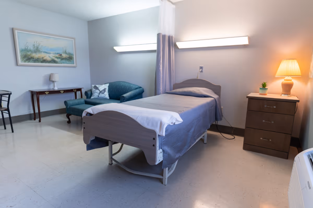 A simple nursing facility room with a single bed covered in gray bedding, a wooden nightstand with a lamp and small plant, a green upholstered loveseat with a decorative pillow, a wooden side table with a small lamp, and a framed landscape painting on the wall.