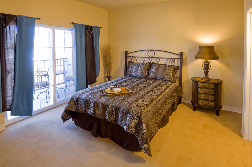 Neatly made bed with patterned bedding in a sunlit bedroom featuring sliding glass doors to a small balcony and a bedside table with a lamp.