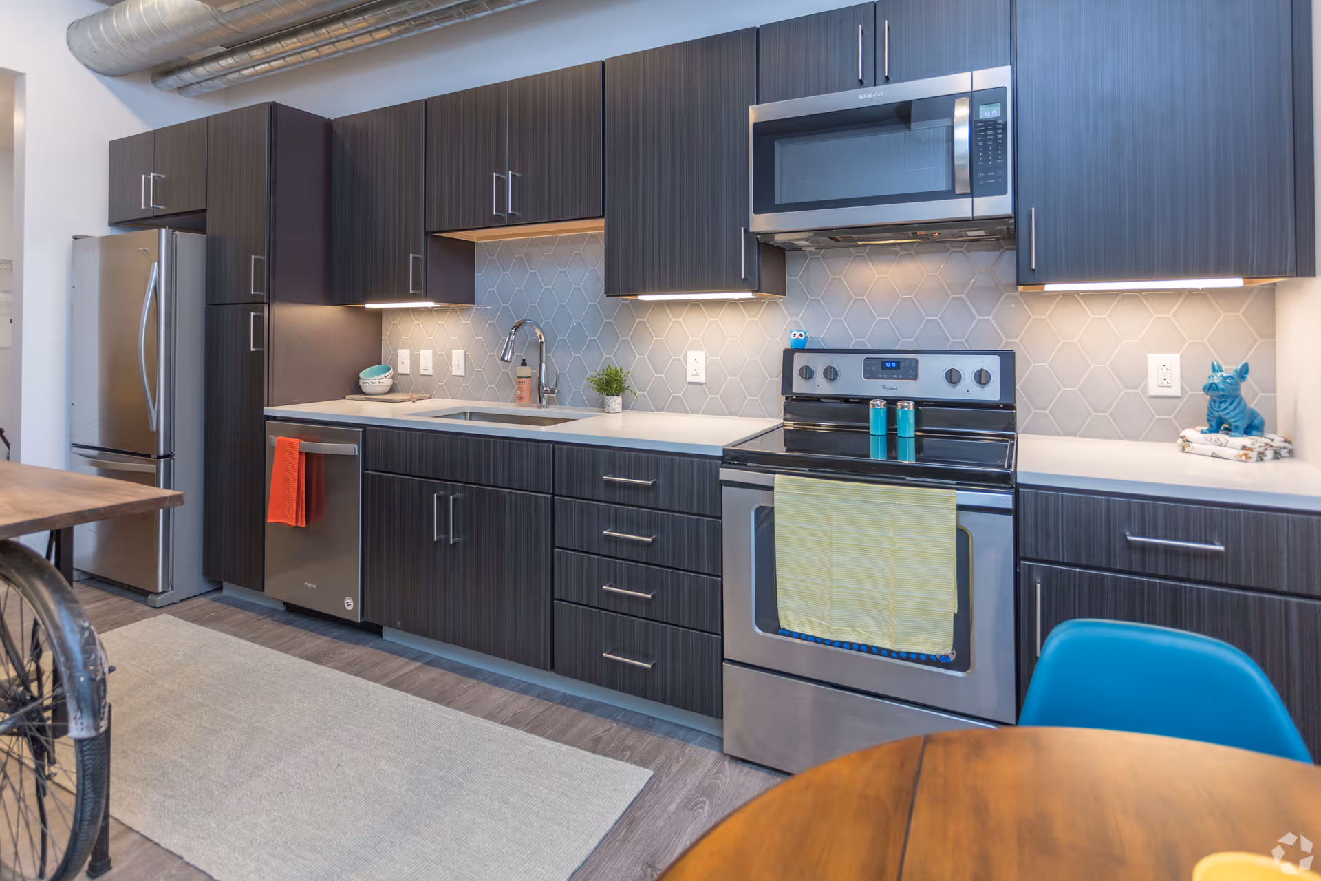 Modern kitchen with dark wood cabinets, stainless steel refrigerator, dishwasher, oven, and microwave. The backsplash features a light gray hexagonal tile pattern. There are small decorative items on the countertop including a small plant, soap dispenser, and a blue dog figurine. A wooden dining table with a blue chair is partially visible in the foreground.