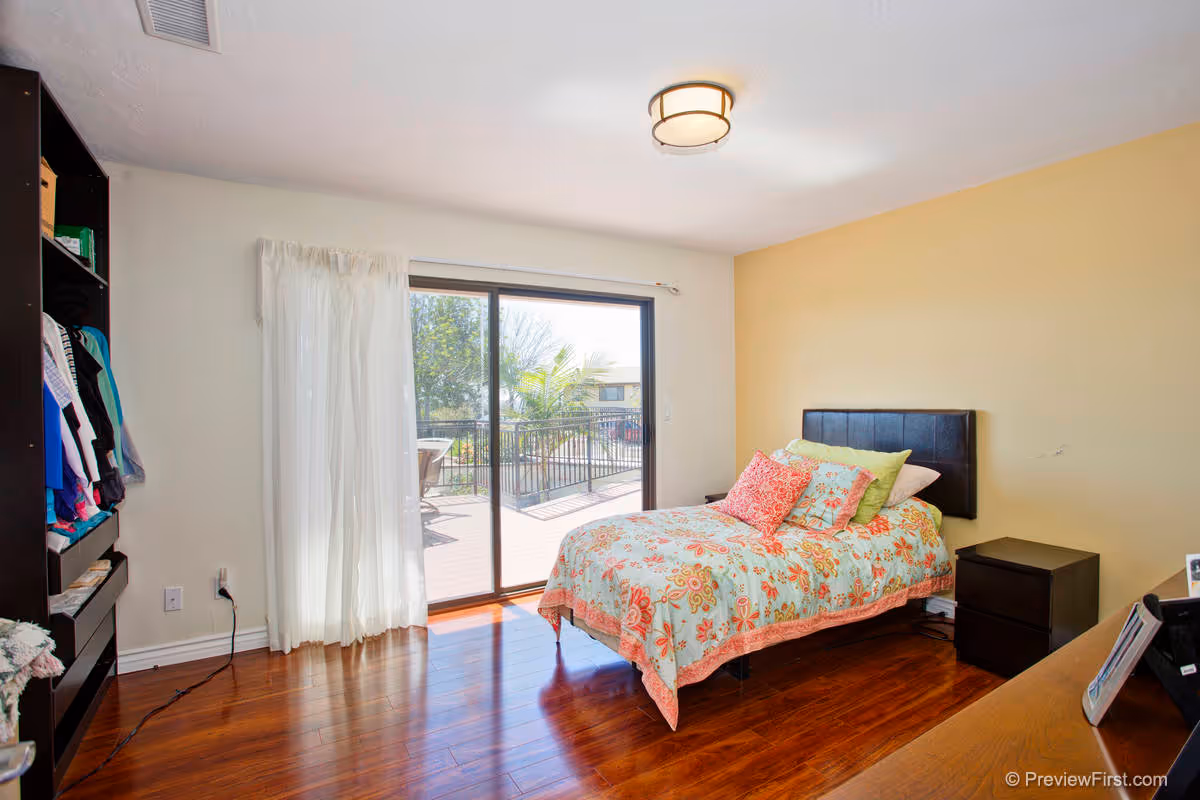 A bright bedroom with a single bed covered in a floral patterned bedspread and multiple pillows. The room has wooden flooring, a black headboard, a dark nightstand, and a dresser. There is a sliding glass door with sheer white curtains leading to an outdoor patio with a table and chairs. A ceiling light fixture is mounted above.