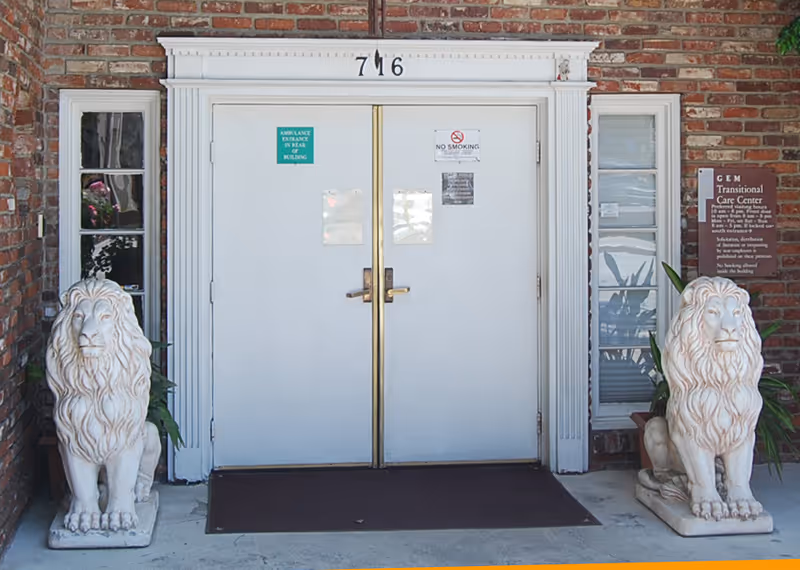 Double white entrance doors with the number 716 above them, flanked by two white lion statues on either side. The building has a brick exterior, and there is a sign on the right side that reads GEM Transitional Care Center with visitor and smoking information.