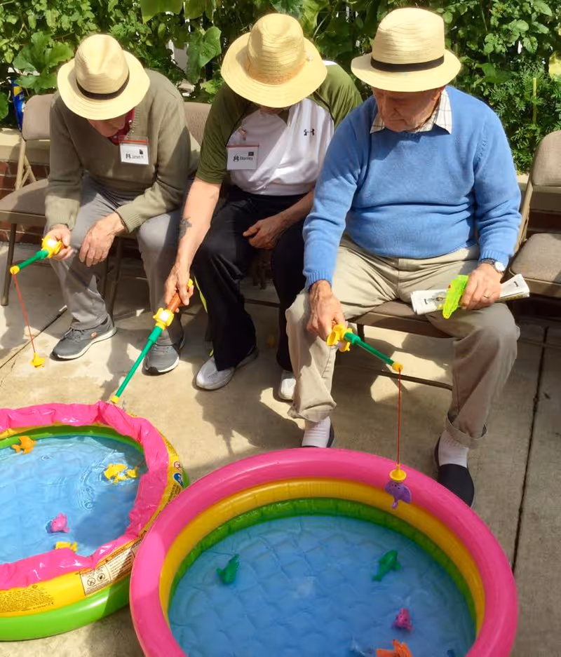 Three elderly individuals wearing straw hats sitting on chairs outdoors, playing a fishing game with small inflatable pools filled with water and floating toy fish.