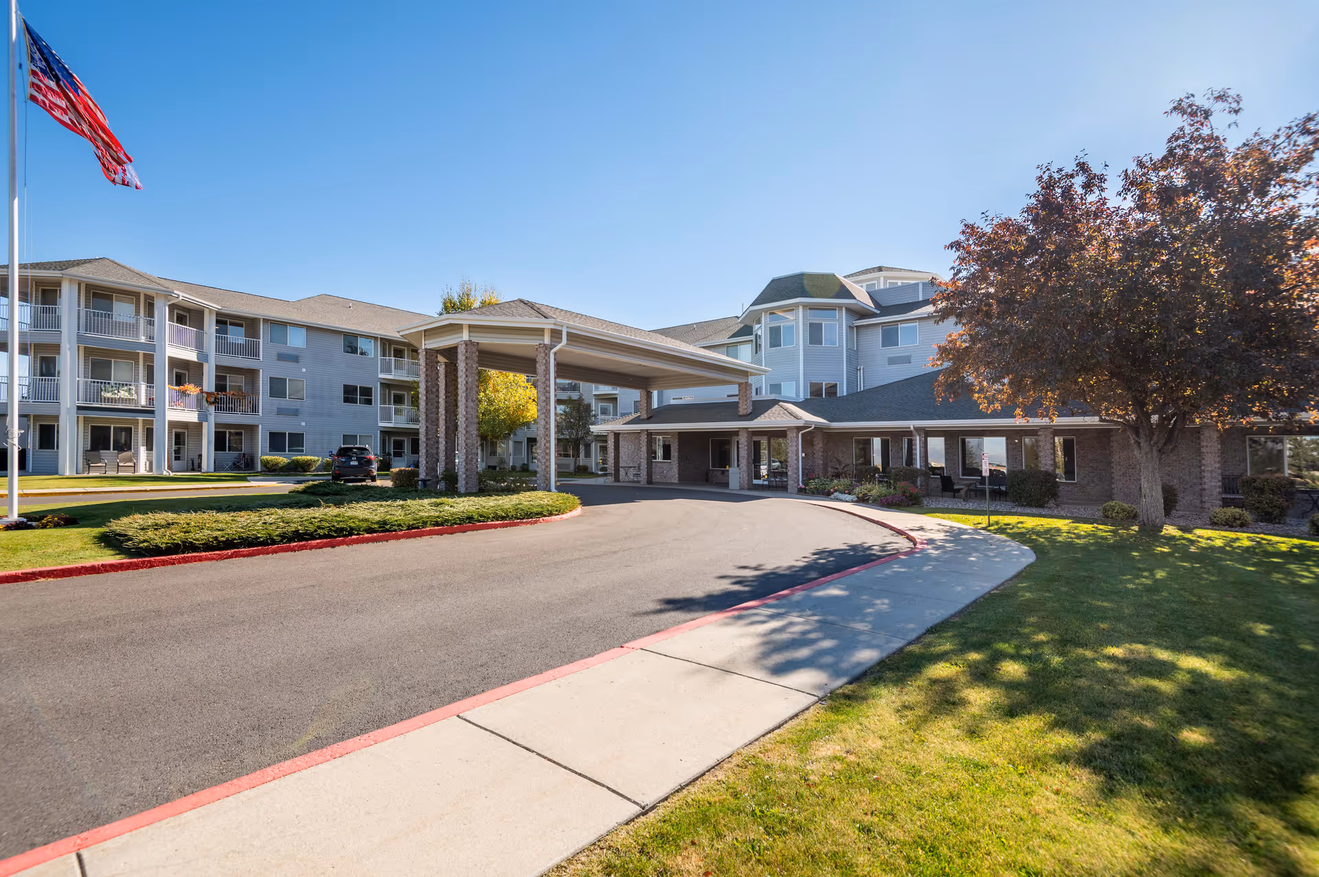 Front exterior of a senior living building with a covered porte-cochere, circular driveway, American flag, and landscaped lawn.