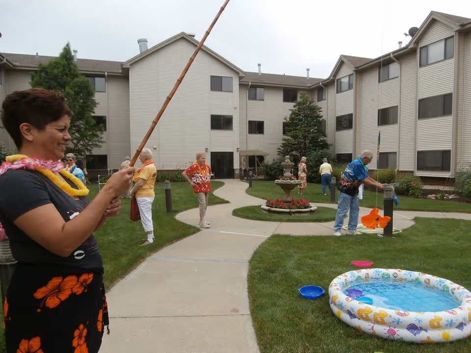 A group of elderly people and a woman are participating in an outdoor fishing game in the courtyard of a multi-story assisted living facility. The woman in the foreground is holding a fishing pole and smiling. The courtyard has a paved walkway, green grass, a small inflatable pool filled with water, and a decorative fountain with flowers in the center. The building surrounding the courtyard has multiple windows and beige siding.