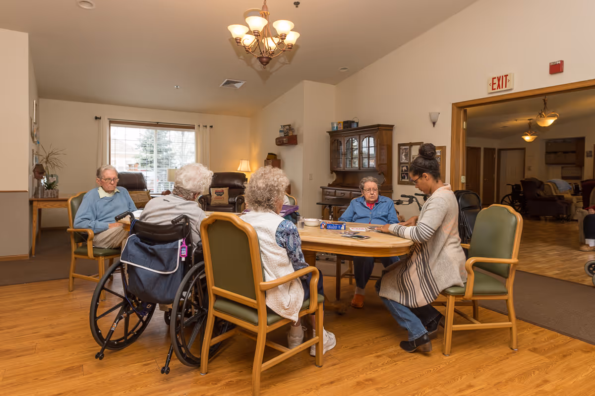 A group of elderly people and a caregiver sitting around a wooden table in a well-lit room with wooden flooring. Some of the elderly individuals are seated in wheelchairs while others are in chairs. The caregiver appears to be engaging with the group, possibly playing a game or doing an activity. The room has a window with curtains, a wooden cabinet, and a chandelier hanging from the ceiling.