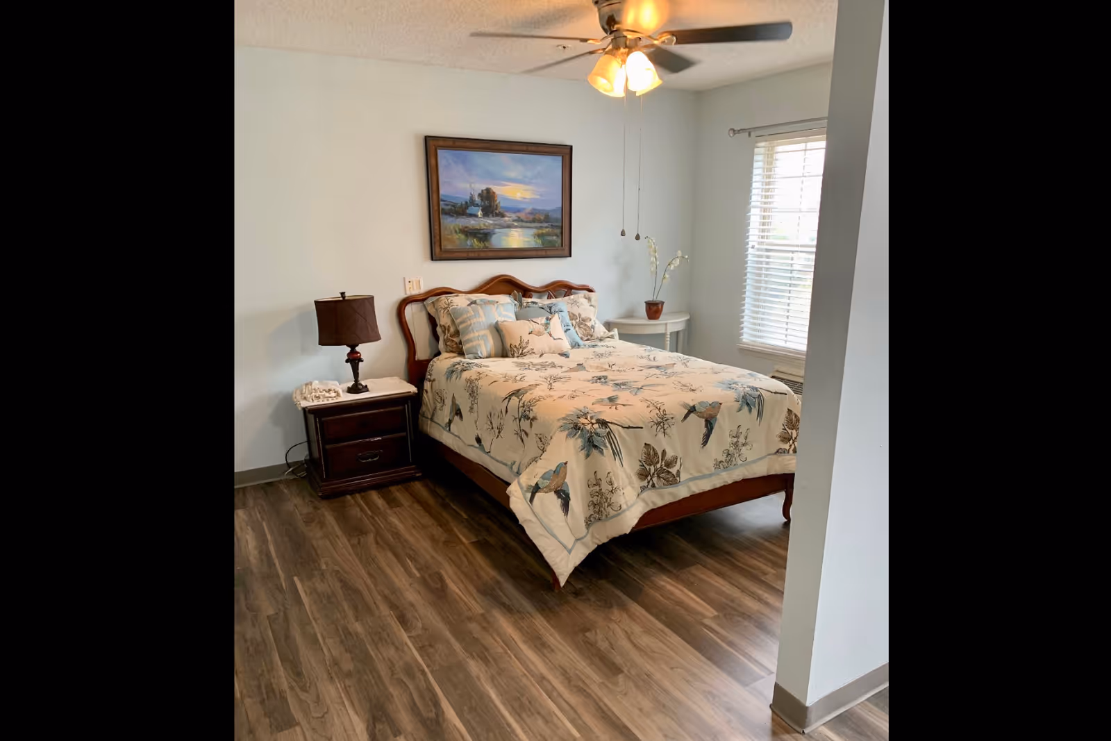 Bedroom with a wooden bed dressed in bird-and-floral bedding, a nightstand with a lamp, ceiling fan, and a window with blinds.