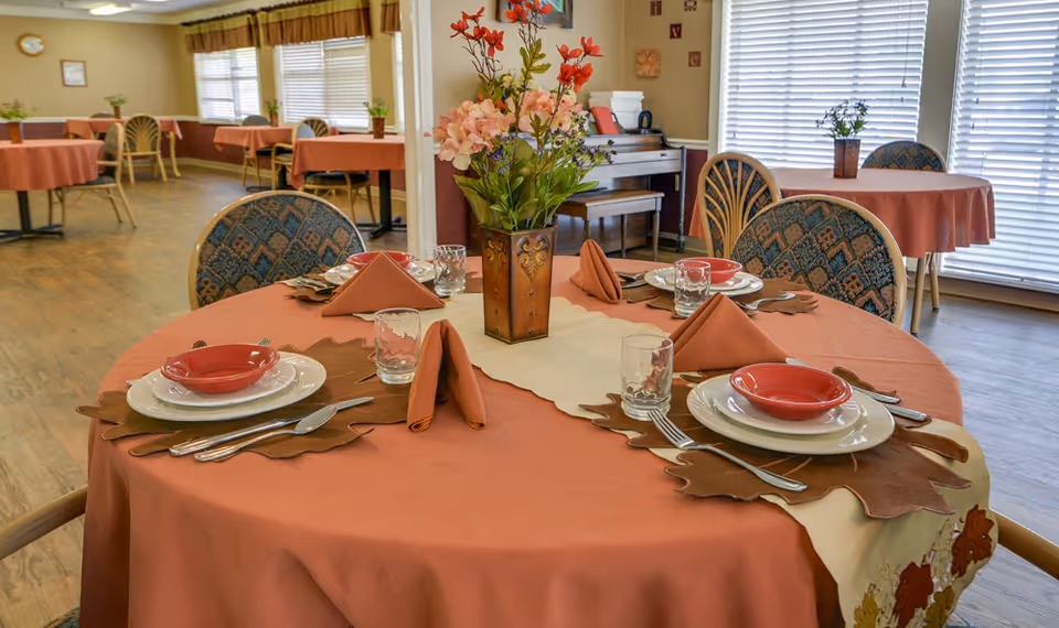 Round dining table set with orange tablecloth, place settings, folded napkins and a floral centerpiece, with additional tables and chairs in the background.
