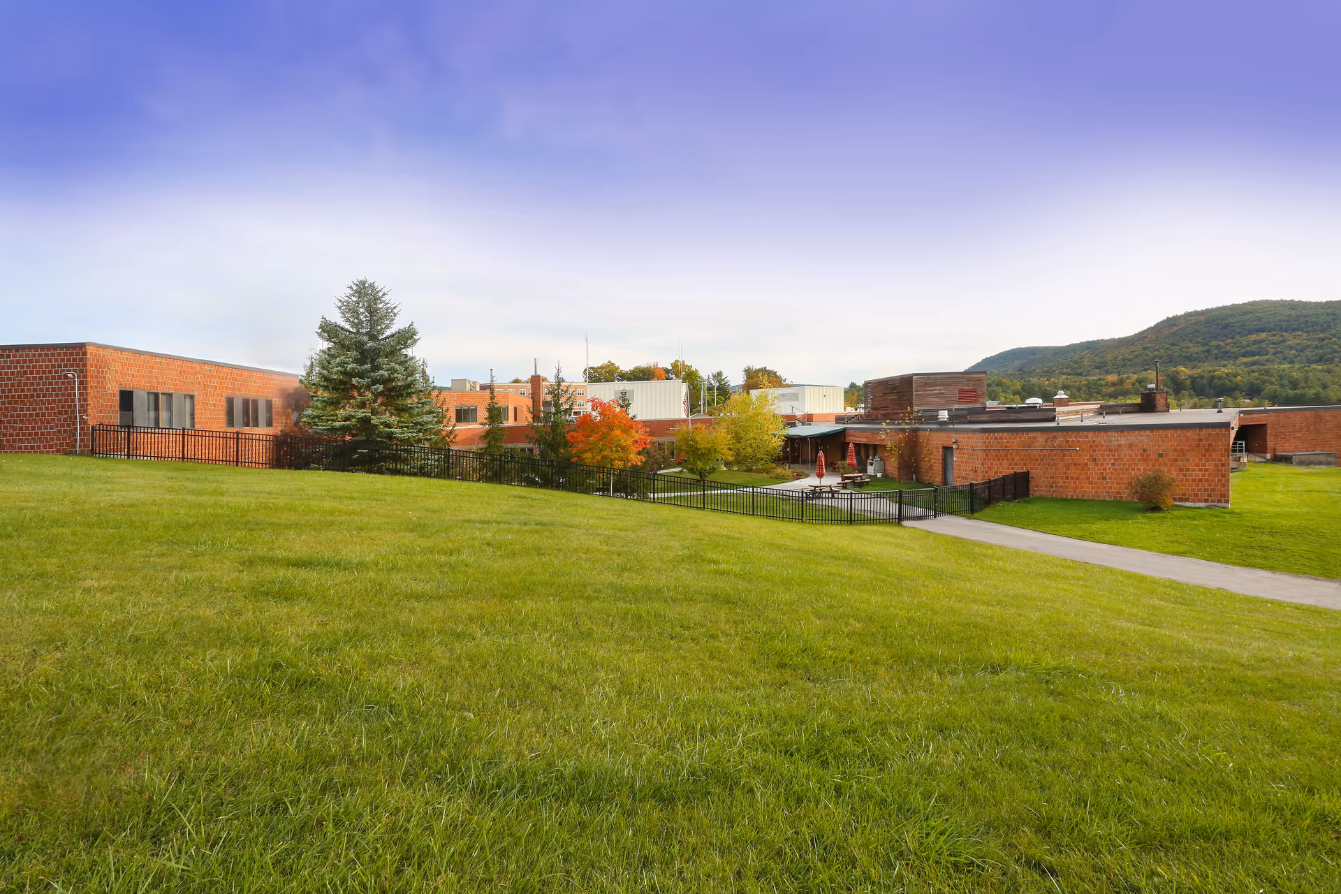 Wide view of Elderwood Village at Ticonderoga showing a large grassy area in the foreground with a paved walkway leading to a low brick building surrounded by trees and a black metal fence, with hills in the background under a clear sky.