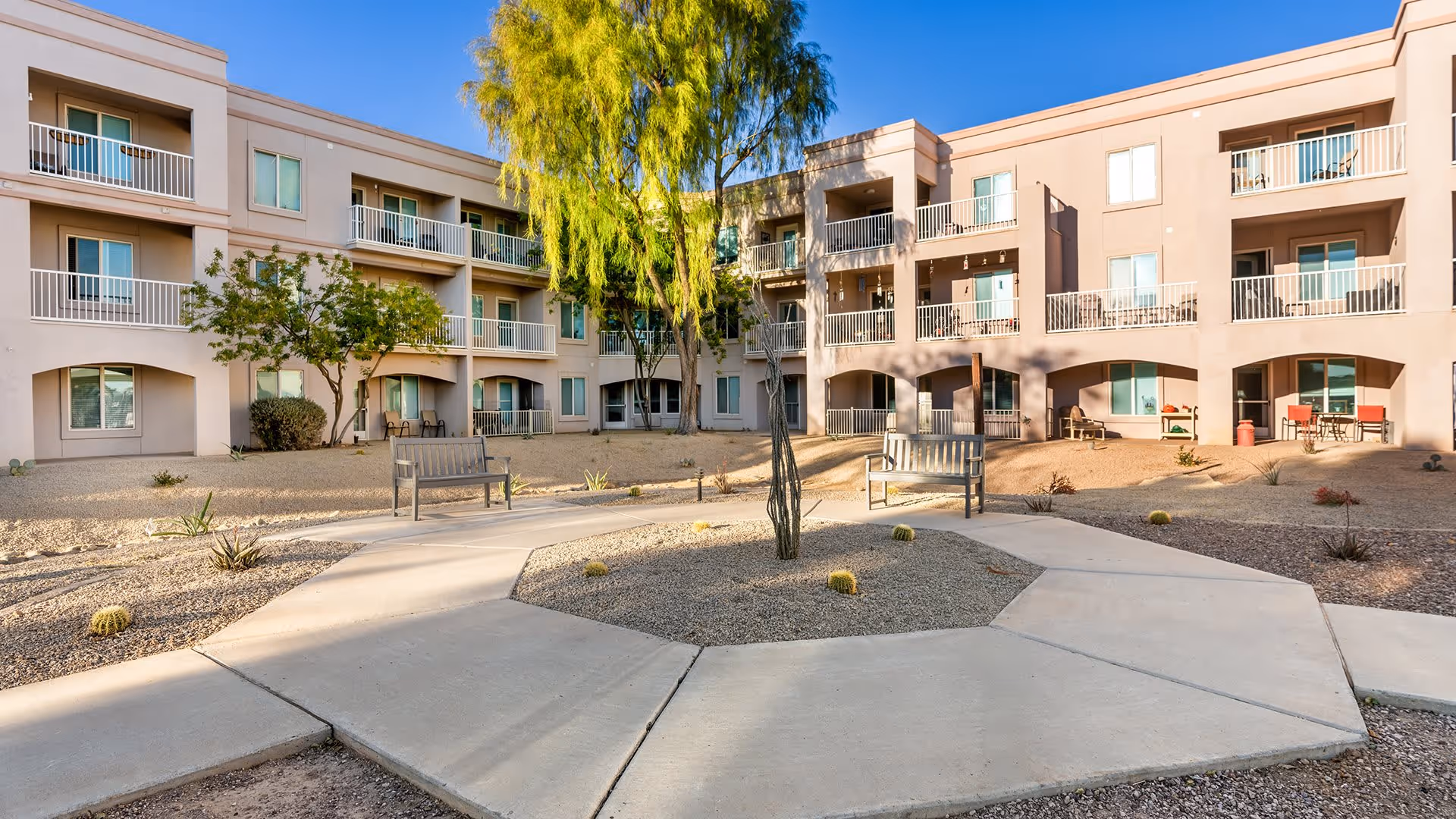 Outdoor courtyard area of a senior living facility with a circular concrete pathway, benches, desert landscaping including small cacti and trees, and a three-story building with balconies and windows surrounding the courtyard.