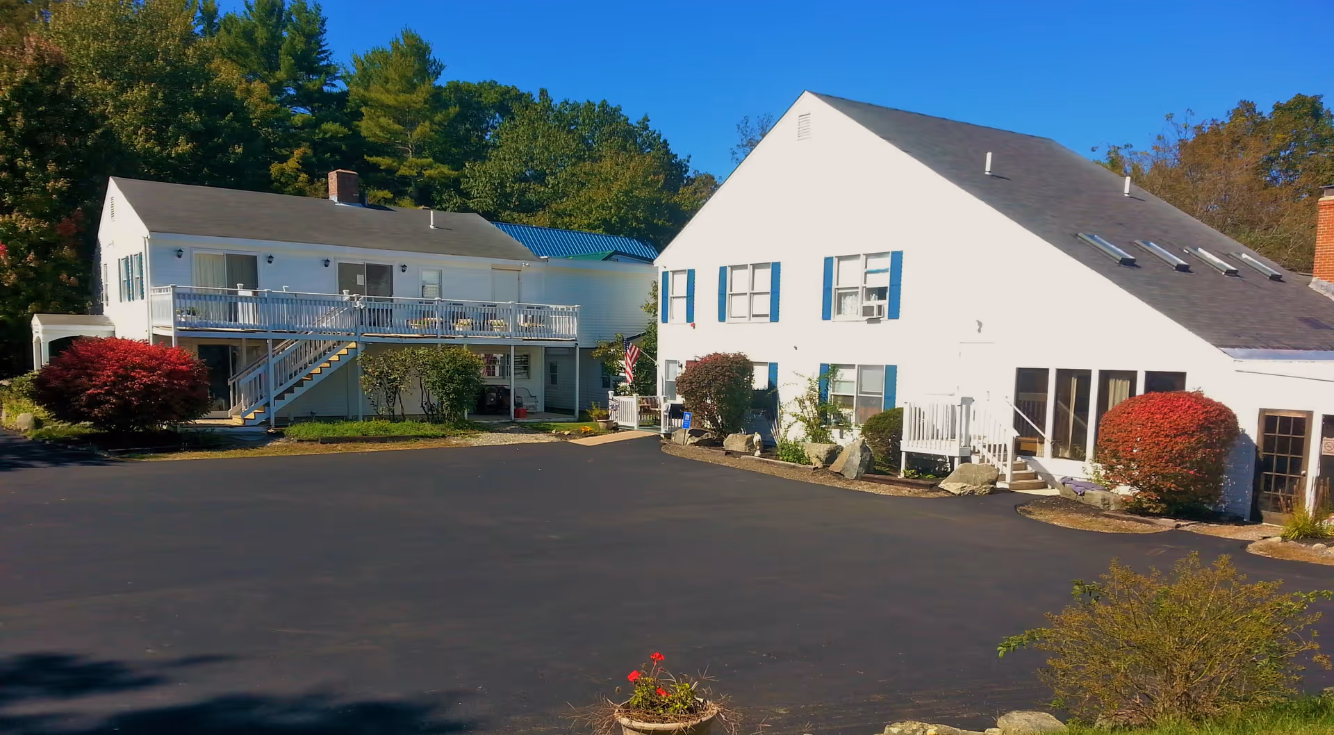 Exterior view of a senior living facility with two white buildings featuring blue shutters, surrounded by greenery and bushes, under a clear blue sky.