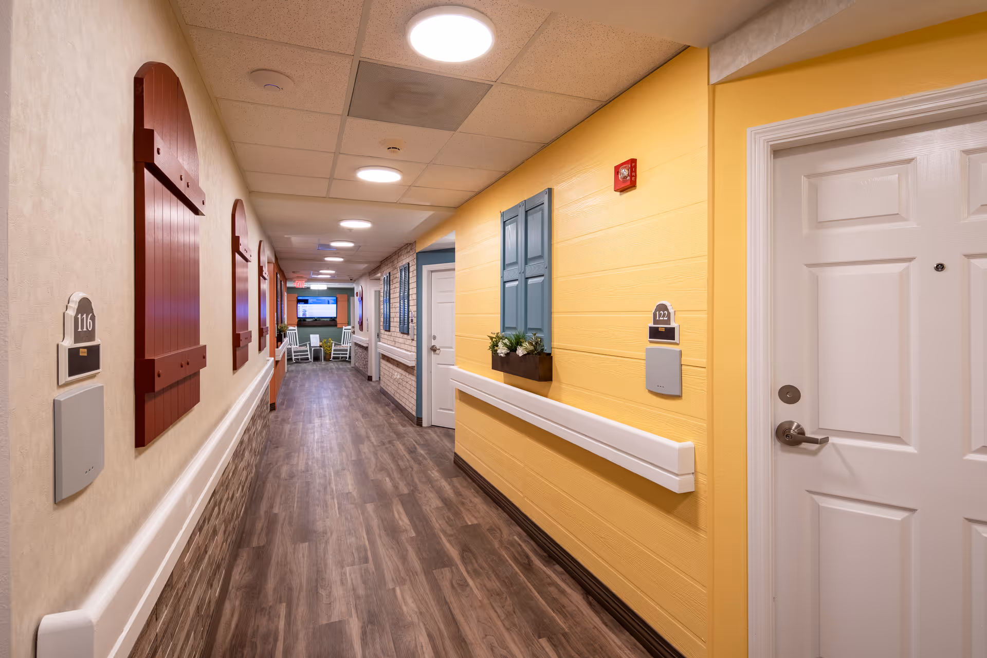 A brightly lit hallway in a senior living facility with wood-style flooring, yellow and beige walls, white doors with room numbers 116 and 122, decorative window shutters, and a seating area with rocking chairs and a television at the far end.