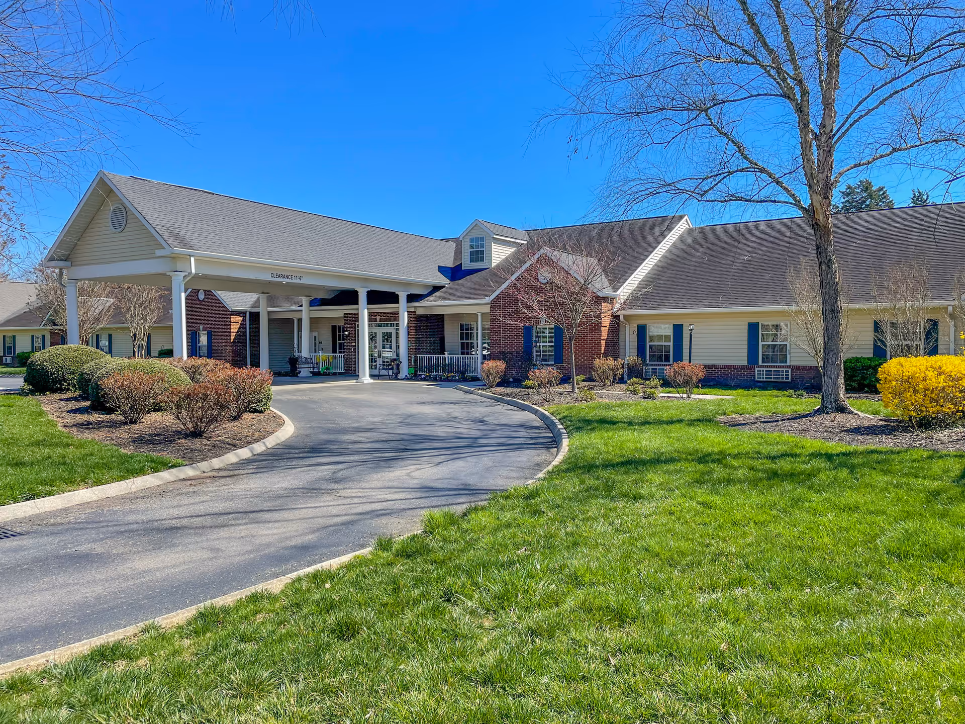 Front entrance of a single-story senior living building with a covered porte-cochere, curved driveway and landscaped lawn.