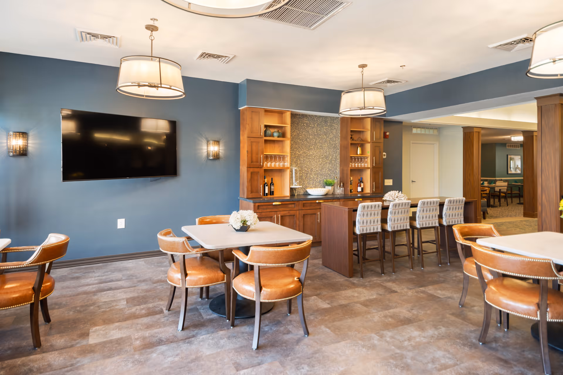 A modern common area with a blue accent wall featuring a large flat-screen TV, two round pendant lights, and wall sconces. The room has several tables with brown leather chairs and a bar area with wooden cabinetry, shelves holding glassware and bottles, and four upholstered bar stools. The flooring is a light brown tile, and there is an open doorway leading to another room with more seating.