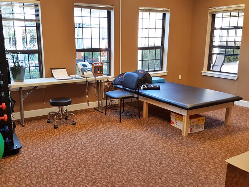 Small therapy/treatment room with a padded examination table, folding table and chairs, medical equipment, and three windows.