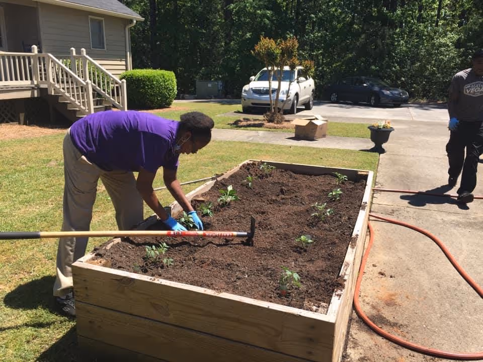 A person wearing a purple shirt and beige pants is gardening in a raised wooden planter box filled with soil and small plants. Another person stands nearby on a concrete driveway with a garden hose. In the background, there is a house with stairs, a lawn, and parked cars surrounded by trees.