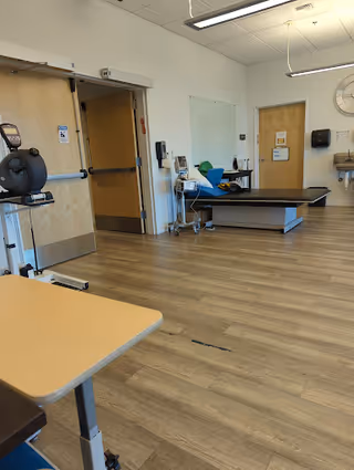 A spacious room with wood-patterned flooring and white walls, featuring a medical examination table, a stationary exercise bike, a small table in the foreground, a wall-mounted clock, and medical equipment near a door. The room appears clean and well-lit with overhead fluorescent lighting.