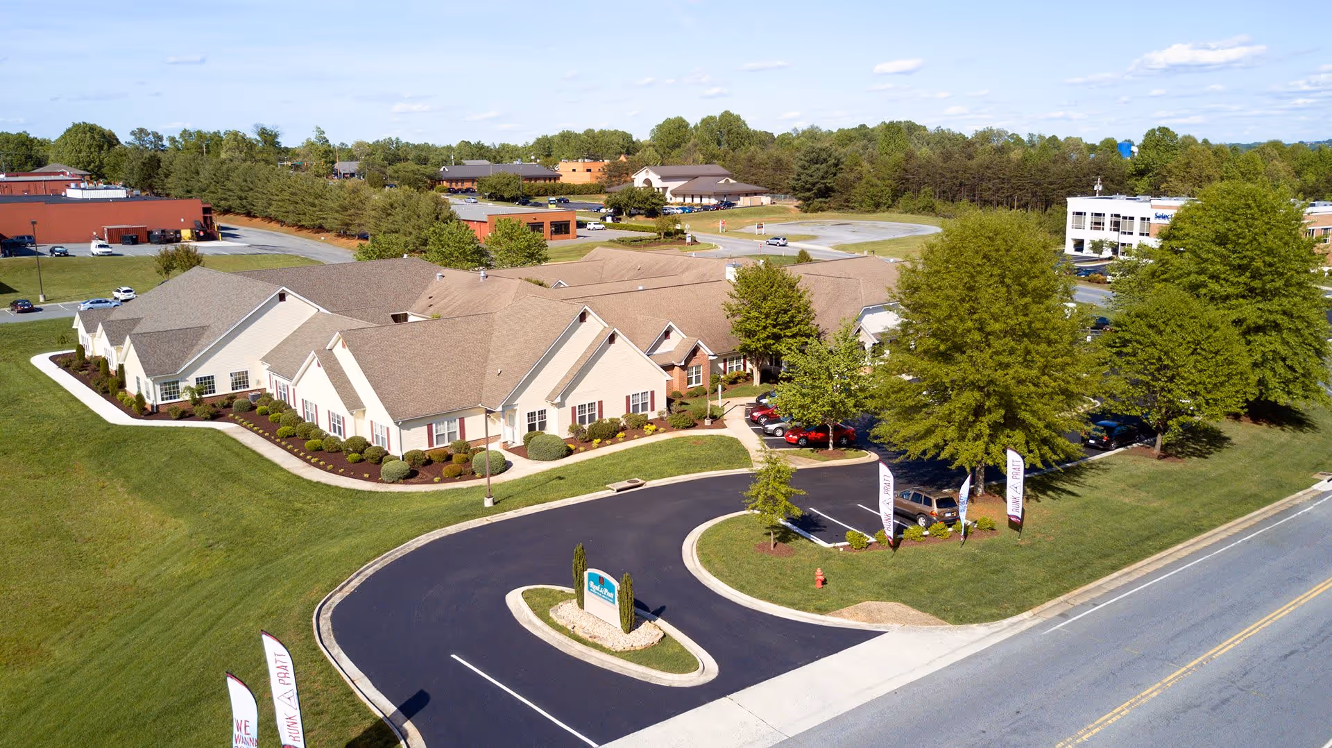 Aerial view of a senior living facility named Runk & Pratt at Forest, showing a large single-story building with beige walls and a brown roof surrounded by well-maintained green lawns, trees, and a curved driveway with a sign at the entrance. Several cars are parked near the building, and other buildings and trees are visible in the background under a partly cloudy sky.