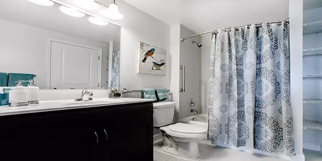 A clean and modern bathroom featuring a dark wood vanity with a white countertop and sink, a large mirror above the sink with three light fixtures, a toilet next to the vanity, and a bathtub with a decorative blue and white patterned shower curtain. There is a towel rack with teal towels and a bird-themed wall art above the toilet. The bathroom has white tiled flooring and light-colored walls.