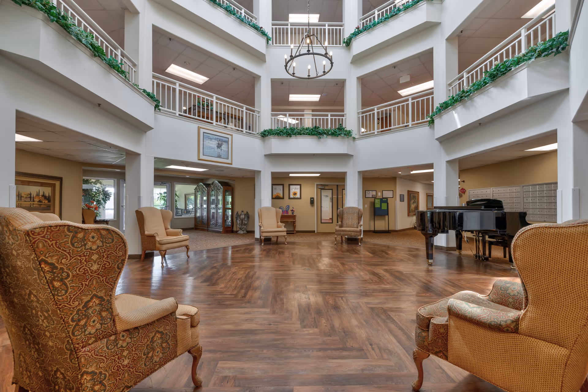 Spacious, multi-level common area in a senior living facility with wooden flooring, several upholstered armchairs arranged around the room, a black grand piano on the right, and white railings with green plants on the upper levels.