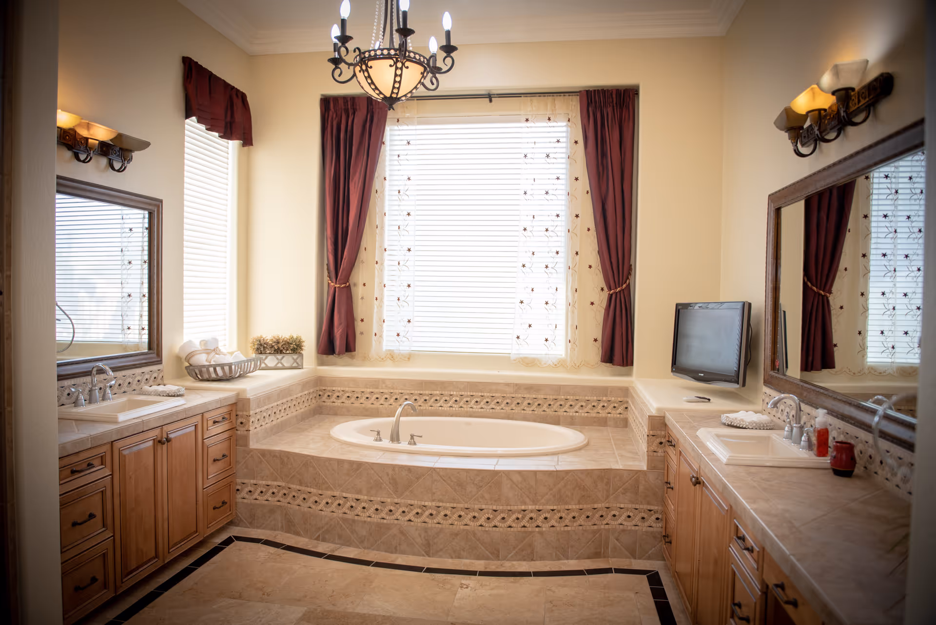 A spacious bathroom with a large built-in bathtub surrounded by beige tiles. The bathtub is centered under a window with white blinds and sheer curtains with a floral pattern, flanked by dark red drapes. On either side of the bathtub are wooden vanities with sinks, mirrors, and wall-mounted light fixtures. A small flat-screen TV is mounted on the right wall above the vanity.