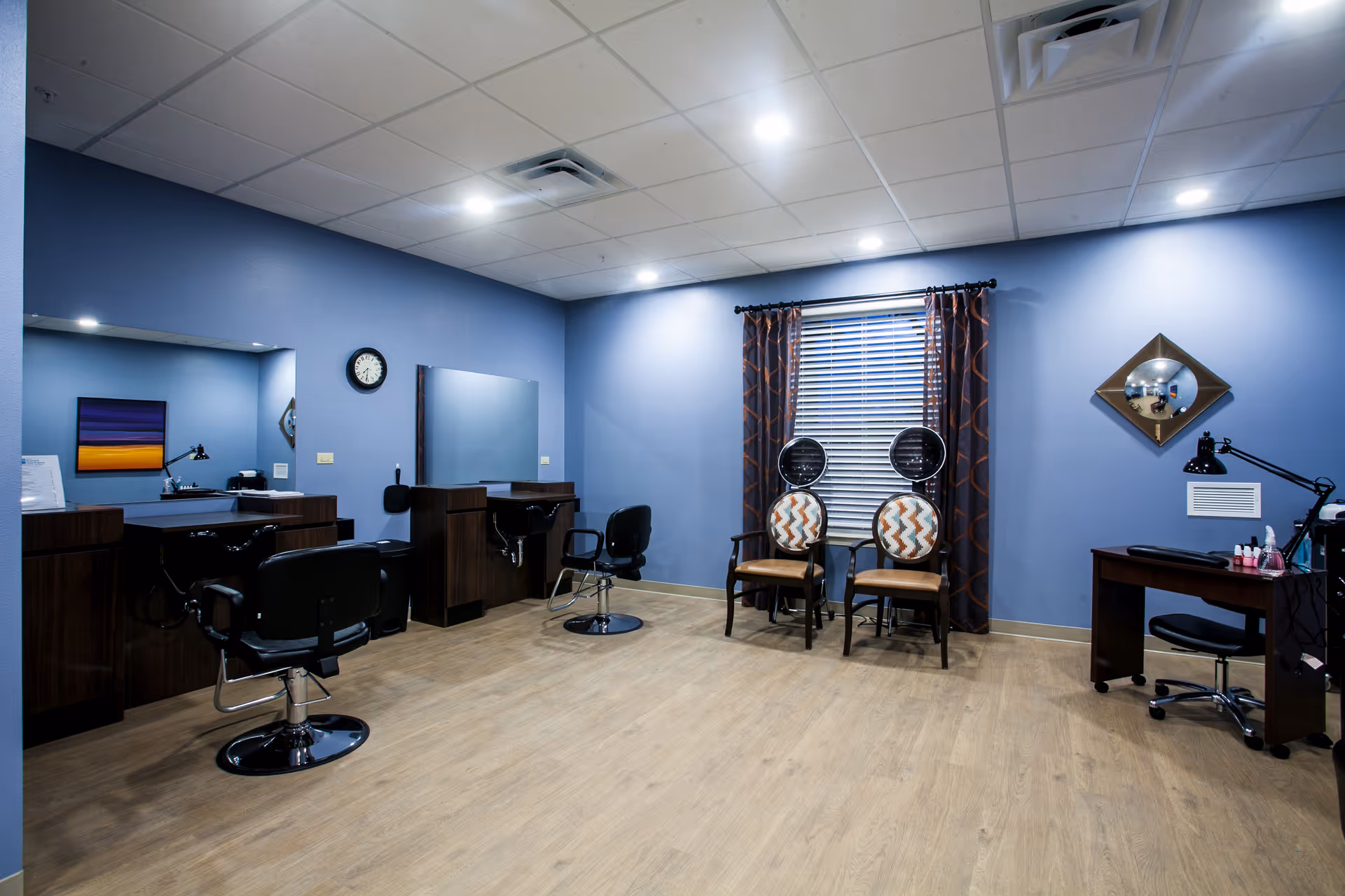 Interior of a salon room with blue walls and wooden flooring. The room features two black salon chairs in front of mirrors and sinks on the left side, two patterned chairs with hair dryers in front of a window with curtains in the center, and a manicure station with a desk, lamp, and chair on the right side.