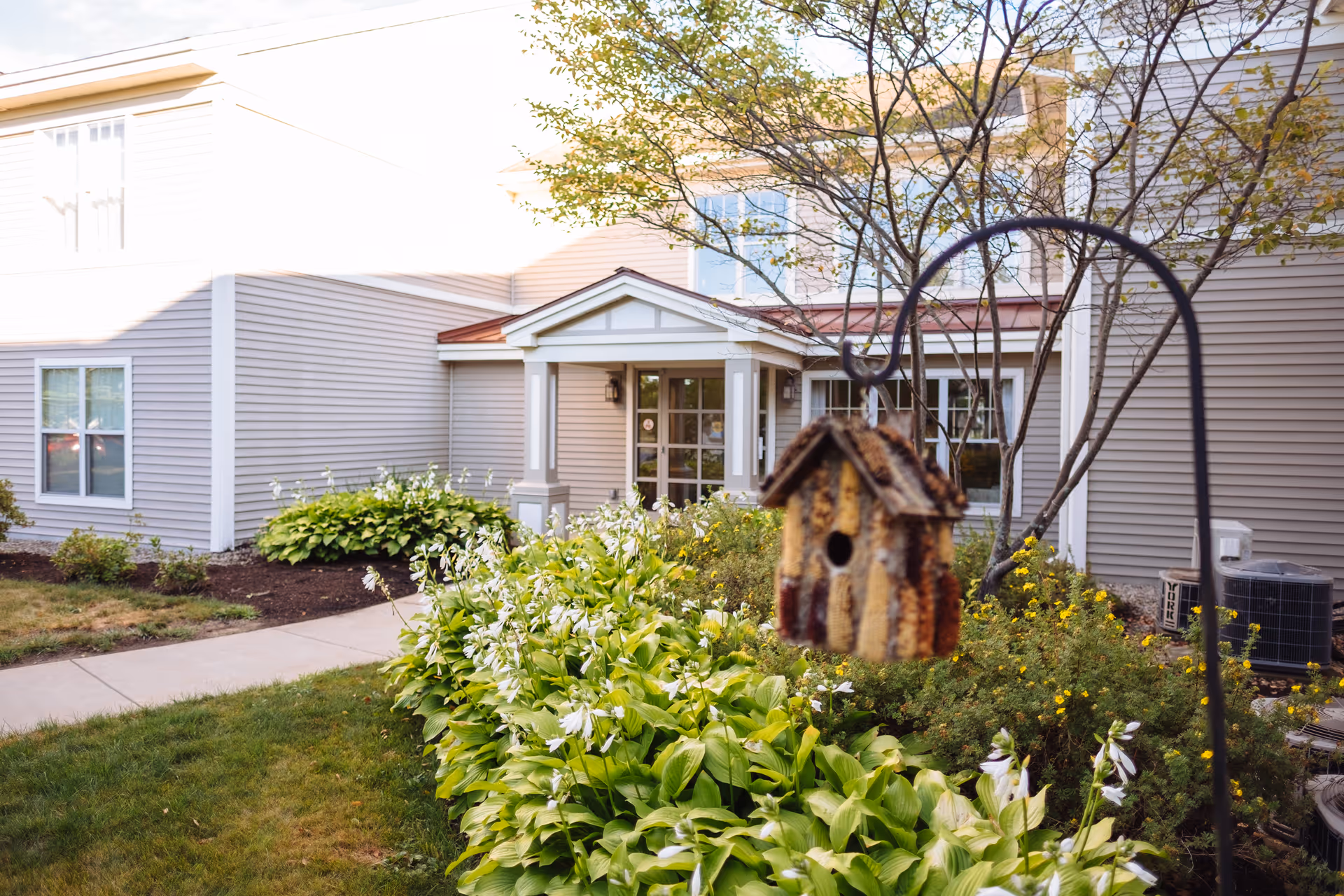 View of the exterior of a senior living facility building with beige siding, white trim, and a small covered entrance. In the foreground, there is a garden with green plants and flowers, and a bird feeder hanging from a metal hook.