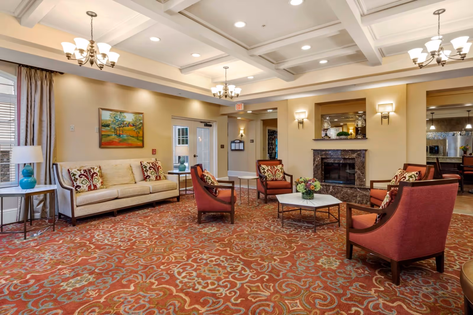 A spacious and well-lit living room area in Symphony Square featuring a beige sofa with patterned cushions, four red armchairs arranged around a white coffee table with a floral centerpiece, a fireplace with a marble surround, and elegant chandeliers hanging from a coffered ceiling. The room has a large patterned carpet and windows with curtains allowing natural light.