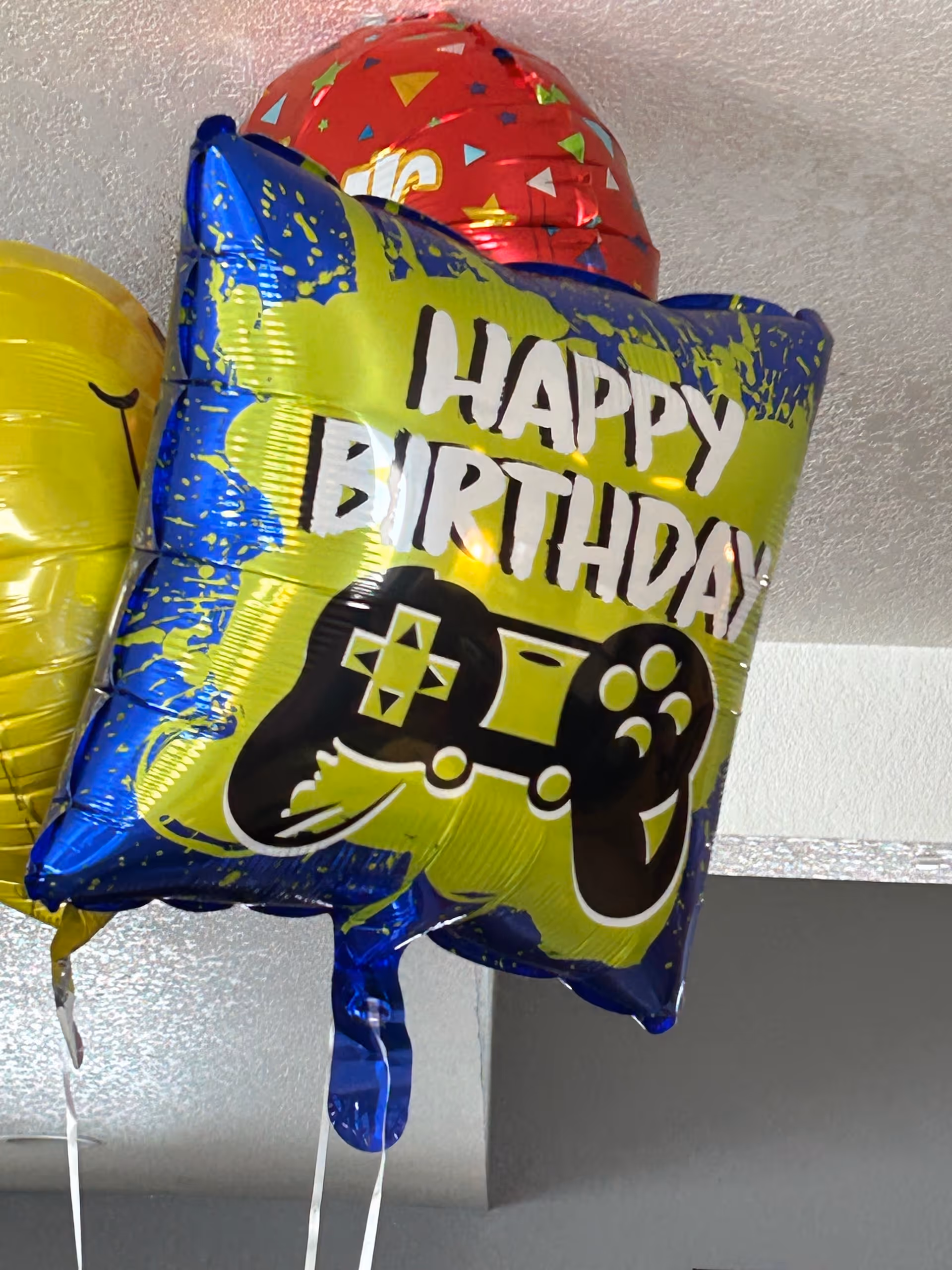 A green-and-blue 'Happy Birthday' mylar balloon featuring a video game controller hanging from a ceiling with other balloons.