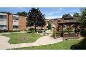 Courtyard of a two-story brick senior living facility with a wooden gazebo, paved walkways, lawn, and trees.
